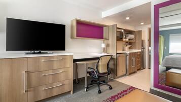 A modern hotel room with a flat-screen TV, a desk with a chair, a kitchenette, and a large mirror reflecting a bed and a bathroom.