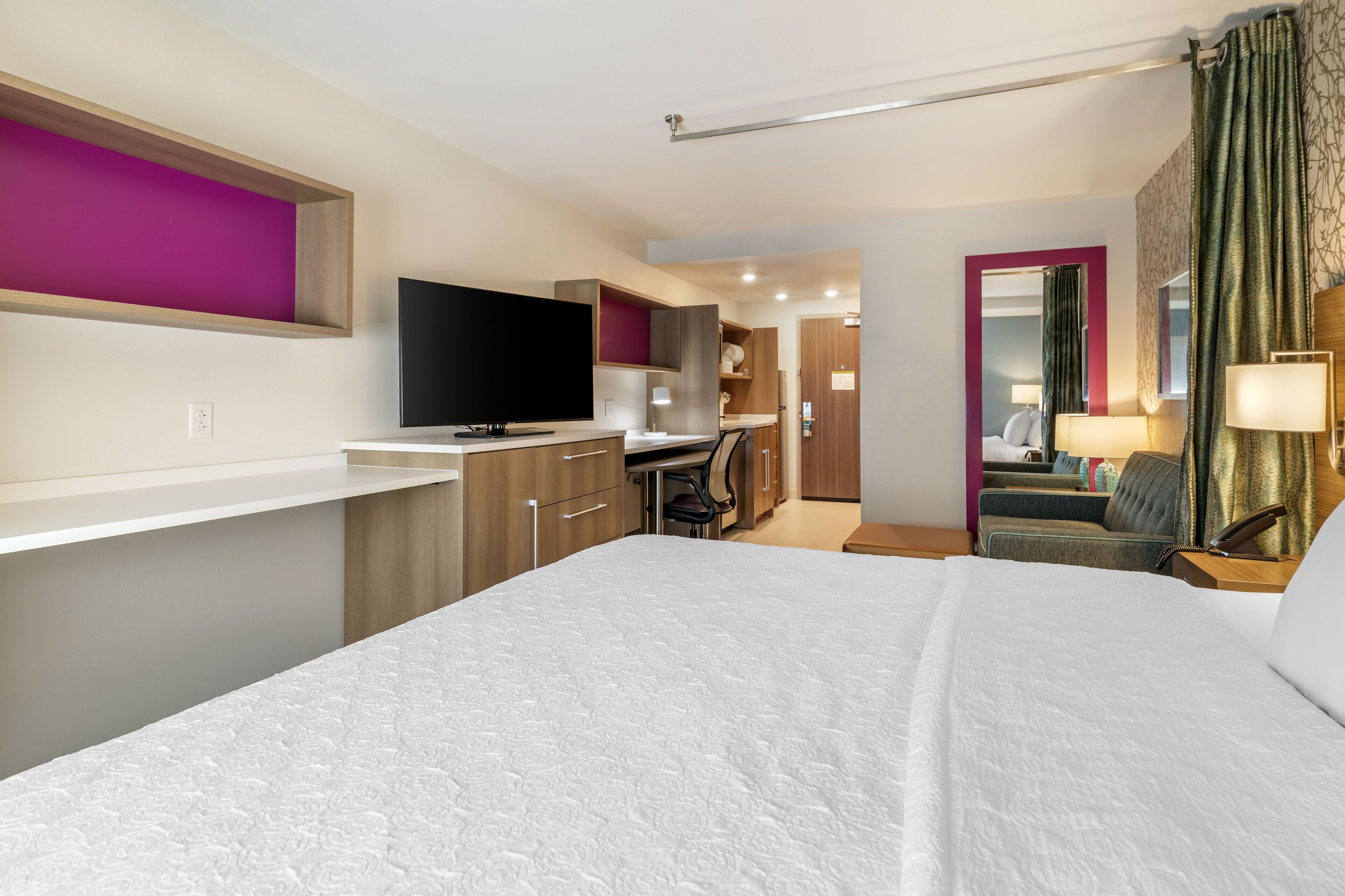 A modern hotel room with a large bed, a flat-screen TV, a desk with a chair, and a separate living area with a sofa and lamp.