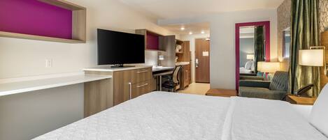 A modern hotel room with a large bed, a flat-screen TV, a desk with a chair, and a separate living area with a sofa and lamp.