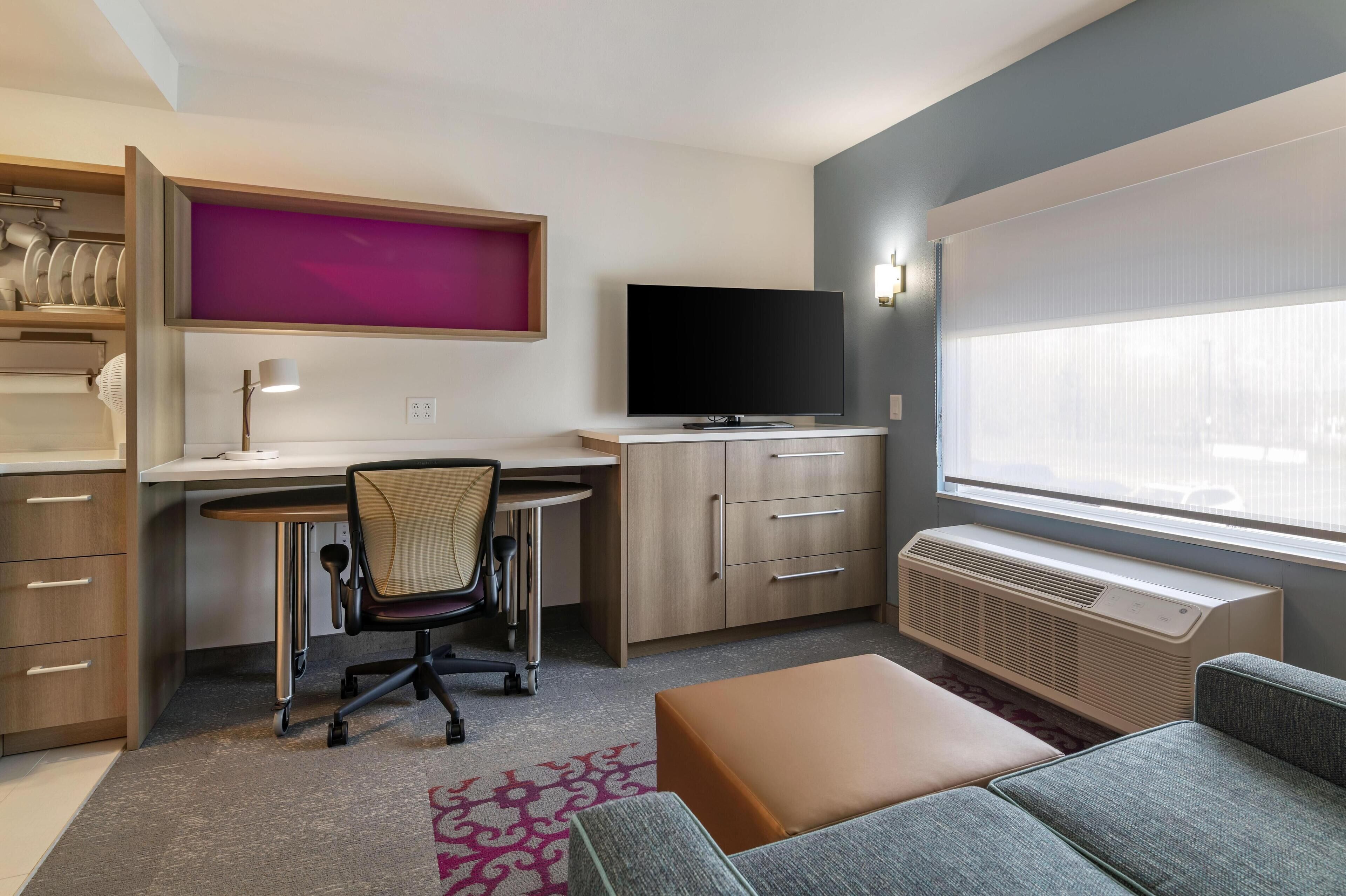 Una habitación de hotel moderna con escritorio, silla, televisión y un ventanal grande.