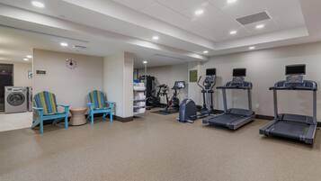 Un gimnasio con equipo de ejercicio, dos sillas azules, una mesita y un reloj en la pared.