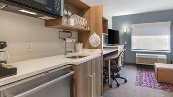 Una cocina moderna con microondas integrado, lavaplatos y armarios. Cuenta con una zona de comedor con escritorio y silla, y un amplio ventanal que proporciona luz natural.
