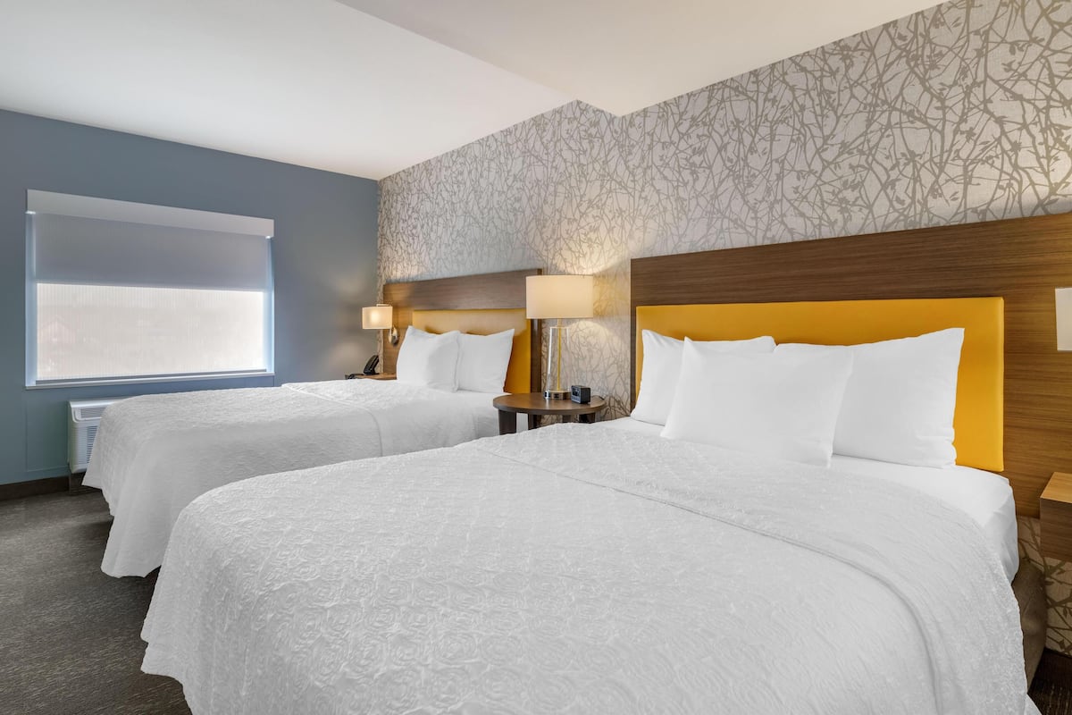 A hotel room with two beds, a large window with blinds, a patterned wall, and a wooden headboard.
