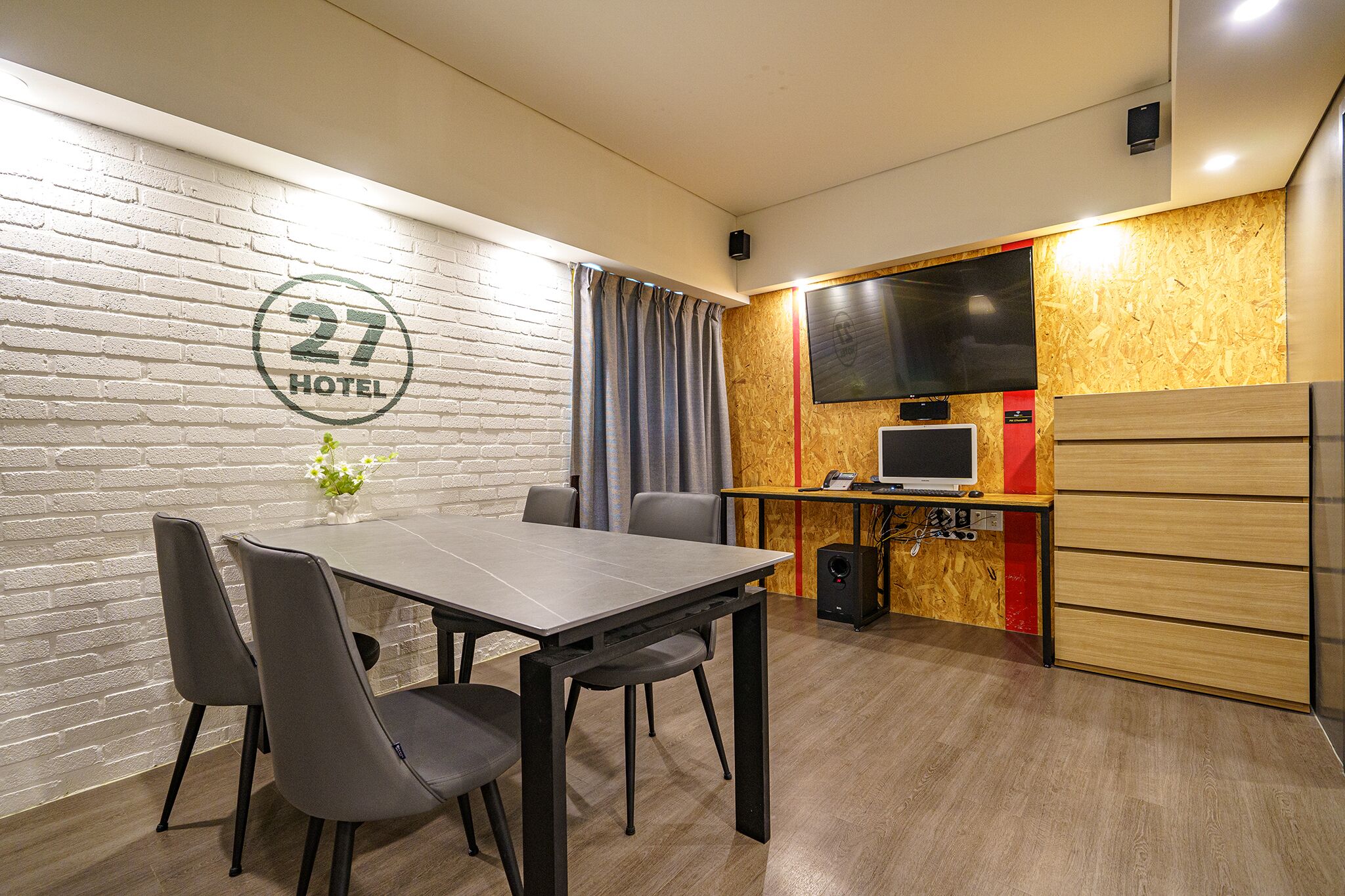 A modern hotel room with a brick wall, a dining table with chairs, a television, and a wooden cabinet.