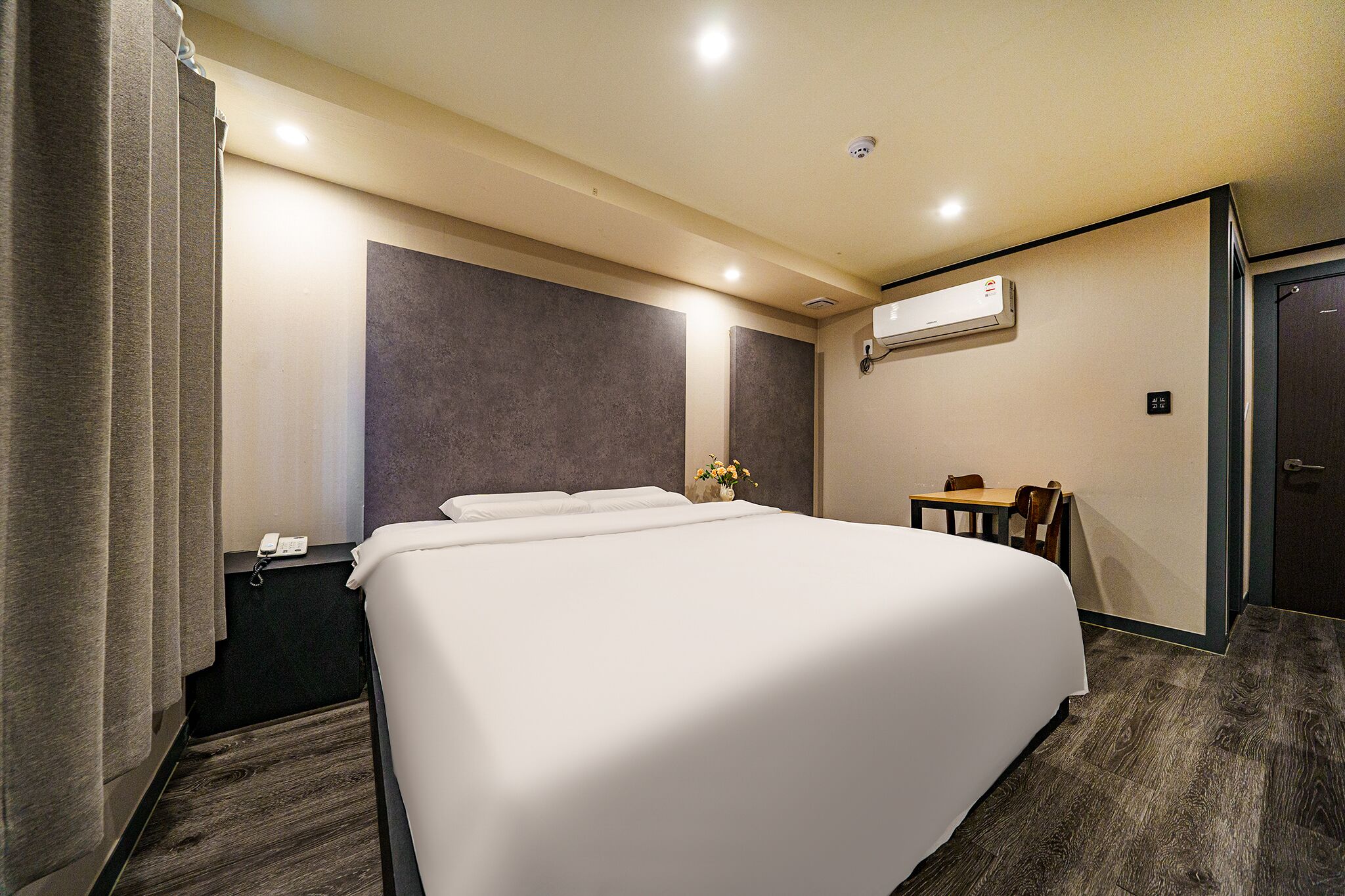 A hotel room with a large bed, a small table, a chair, an air conditioning unit, and a door.