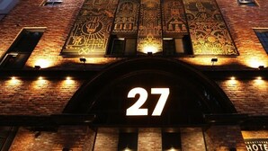 A hotel entrance with a large illuminated number "27" sign, brick facade, and decorative patterns on the upper part of the building.
