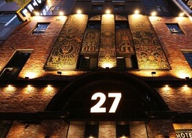 Entrada de hotel com um grande número iluminado "27", fachada de tijolos e padrões decorativos na parte superior do edifício.