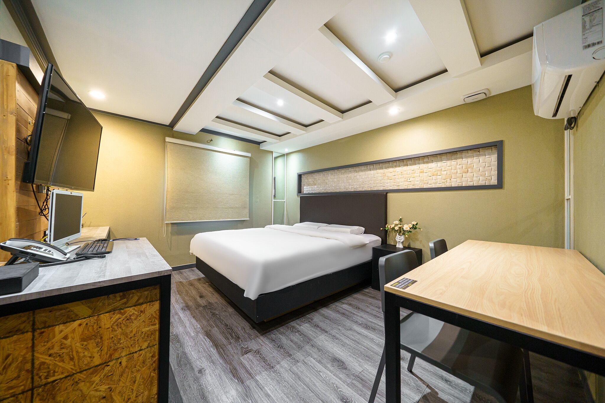 A hotel room with a bed, desk, and chair, a flat-screen TV, a phone, and an air conditioning unit.