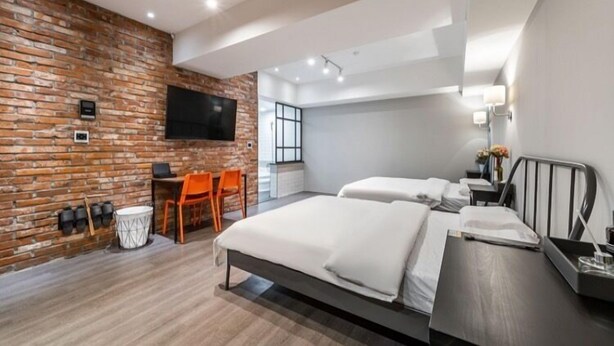 Una habitación de hotel moderna con pared de ladrillo, un televisor de pantalla plana, una cama con sábanas blancas, un sillón naranja y una mesita con lámpara.