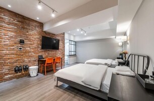 Quarto de hotel moderno com parede de tijolos, televisão de tela plana, cama com lençóis brancos, uma cadeira laranja e uma pequena mesa com abajur.