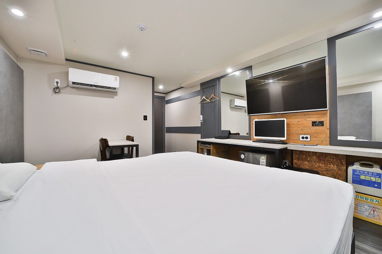 A hotel room with a bed, a desk with a computer, a TV, and an air conditioning unit.