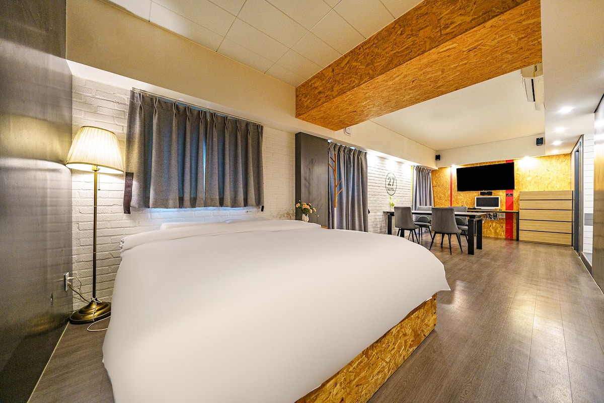 Quarto de hotel com uma cama grande, uma escrivaninha com cadeiras, uma televisão e uma kitchenette.