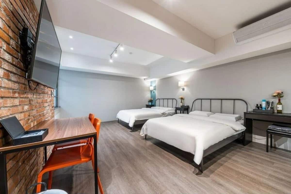 Quarto de hotel moderno com duas camas de solteiro, uma escrivaninha com um laptop, parede de tijolos e uma TV de tela plana na parede.