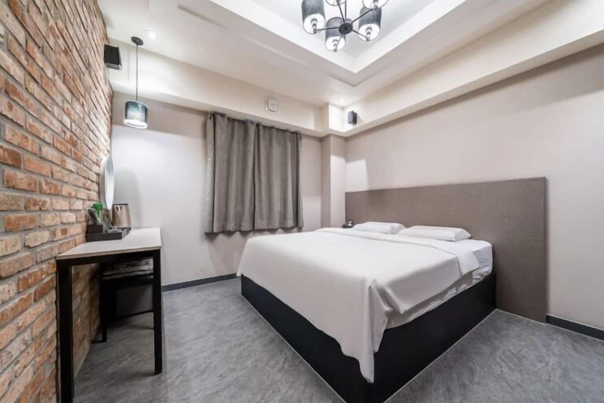 Quarto de hotel moderno com parede de tijolos decorativa, escrivaninha com luminária, cama com roupa de cama branca e claraboia.