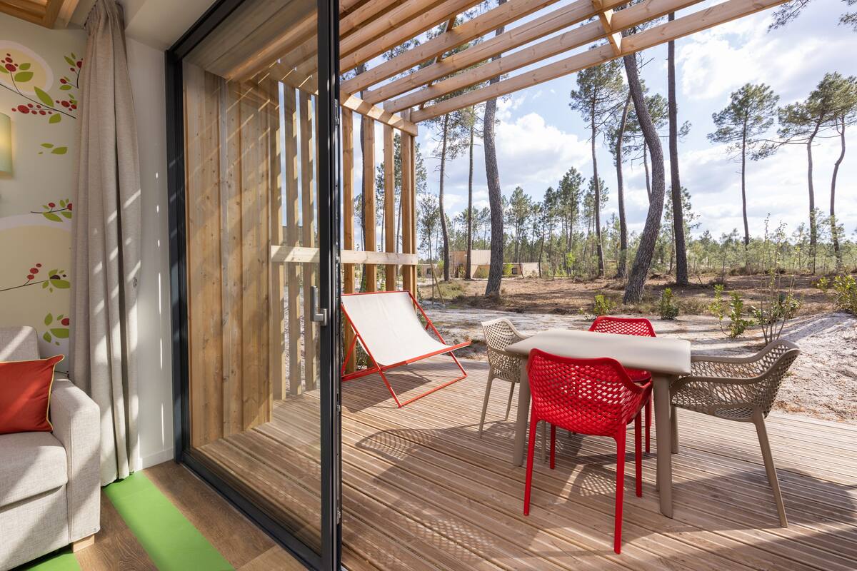 Een moderne kamer met een glazen deur naar een terras. Binnen staat een bank met een rood kussen, een witte stoel en een tafel met rode stoelen. Het terras heeft een houten vloer en uitzicht op een bebost gebied.