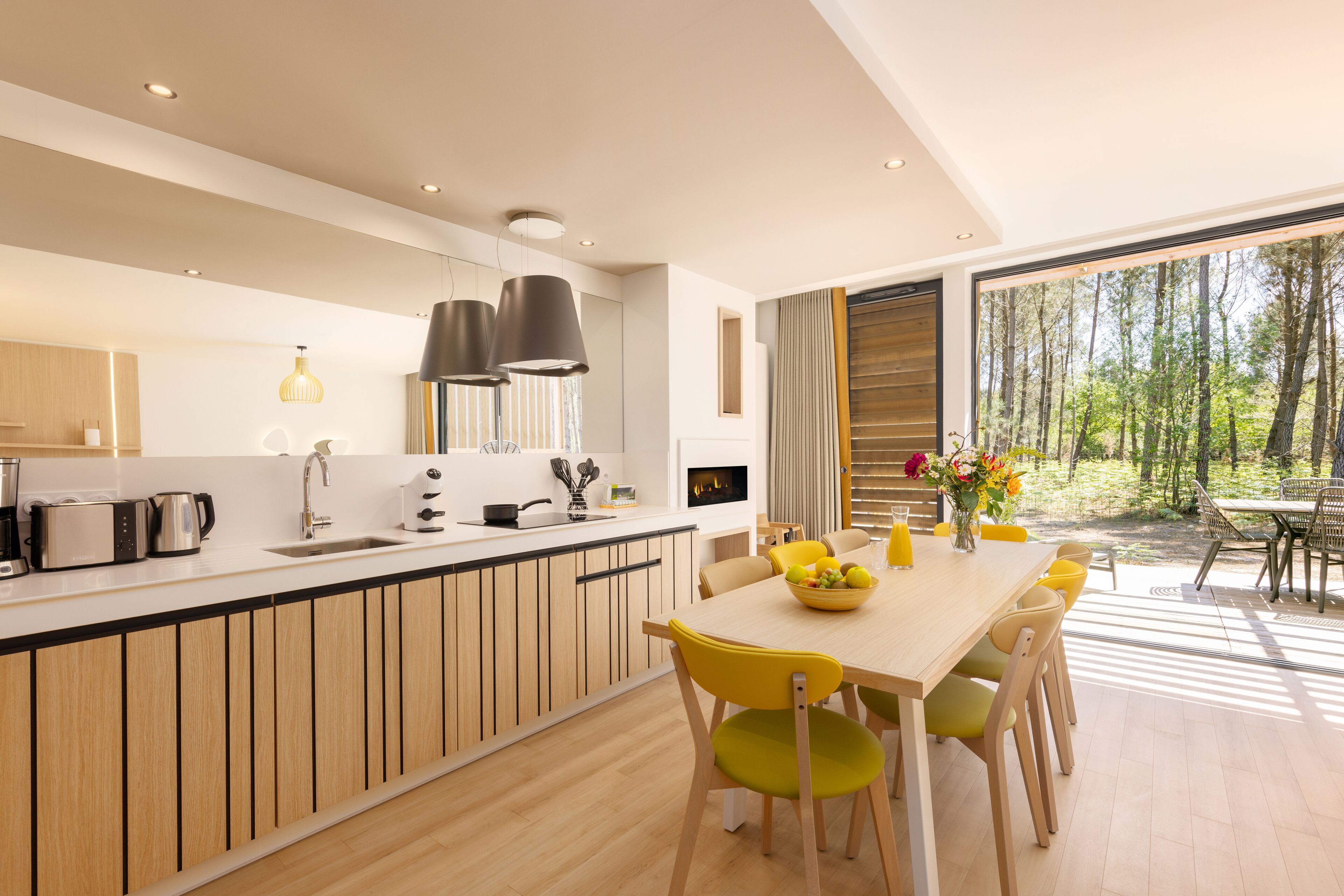 A modern kitchen with a central island, a dining area with yellow chairs, and a view of a forest through large windows.