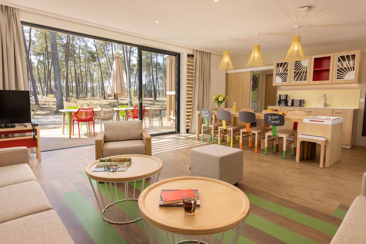 Een moderne hotelkamer met een grote schuifdeur naar uitzicht op een bos, een knus zitgedeelte met een flatscreen televisie, en een kleine kitchenette.