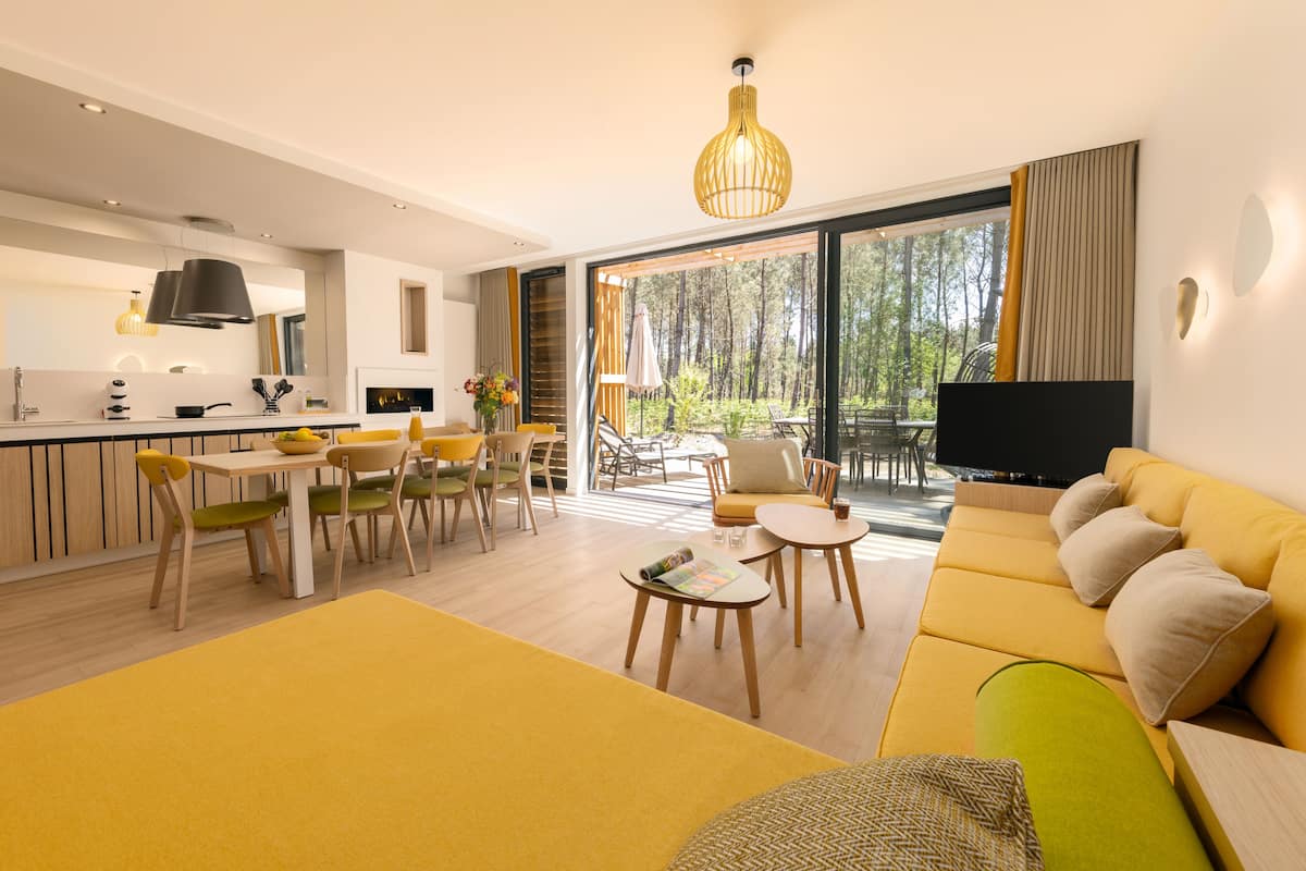 Een moderne woonkamer met een gele bank, een eethoek met houten stoelen, en een keuken met een groot raam met uitzicht op een bos.