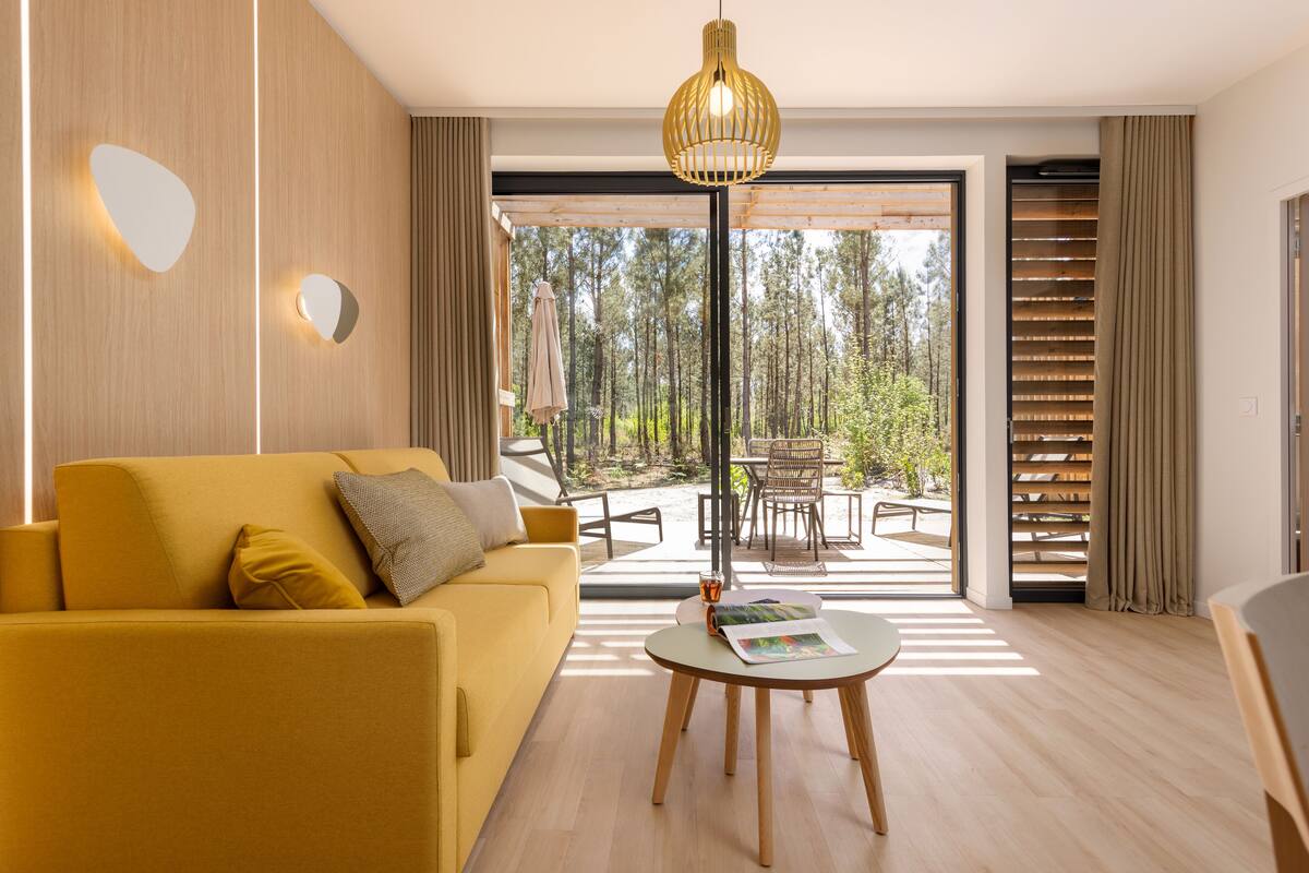 Een moderne woonkamer met een gele bank, een ronde salontafel en een groot raam met uitzicht op een bos.