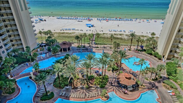 Een resort met een groot zwembad, palmbomen en uitzicht op het strand.