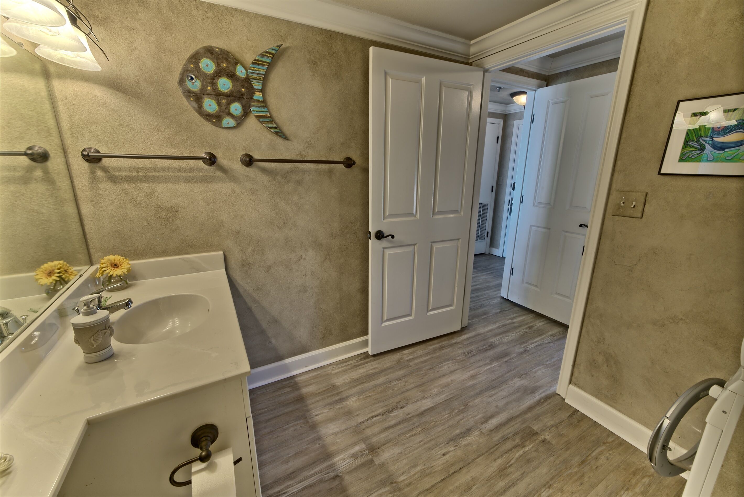 Un baño con lavamanos blanco, un perchero, una puerta y un pasillo que conduce a otra habitación.