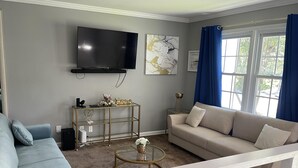 A living room with a beige sofa, a blue armchair, a coffee table, a TV mounted on the wall, and a window with blue curtains.