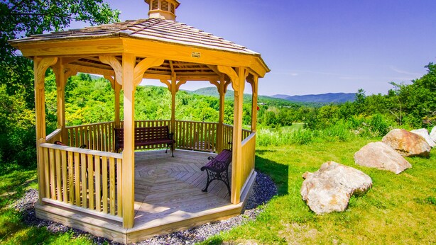 Um gazebo de madeira com um banco e uma pequena torre, rodeado por vegetação exuberante e pedras grandes.