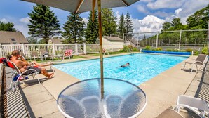 Piscina com espreguiçadeiras e mesa, cercada por grade e árvores.