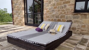 A wicker bed with a grey and white striped bedspread, two pillows, and a rolled-up purple towel on a stone-tiled patio.