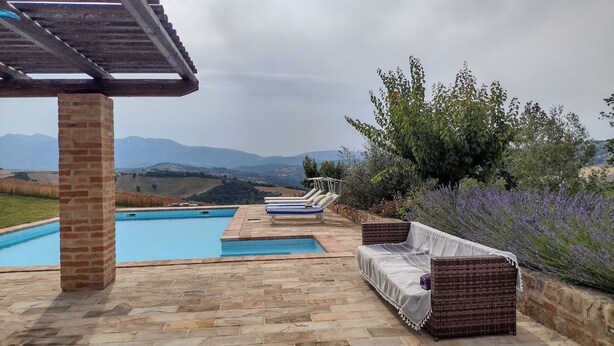 A pool area with a pergola, stone flooring, lounge chairs, and a view of distant mountains.