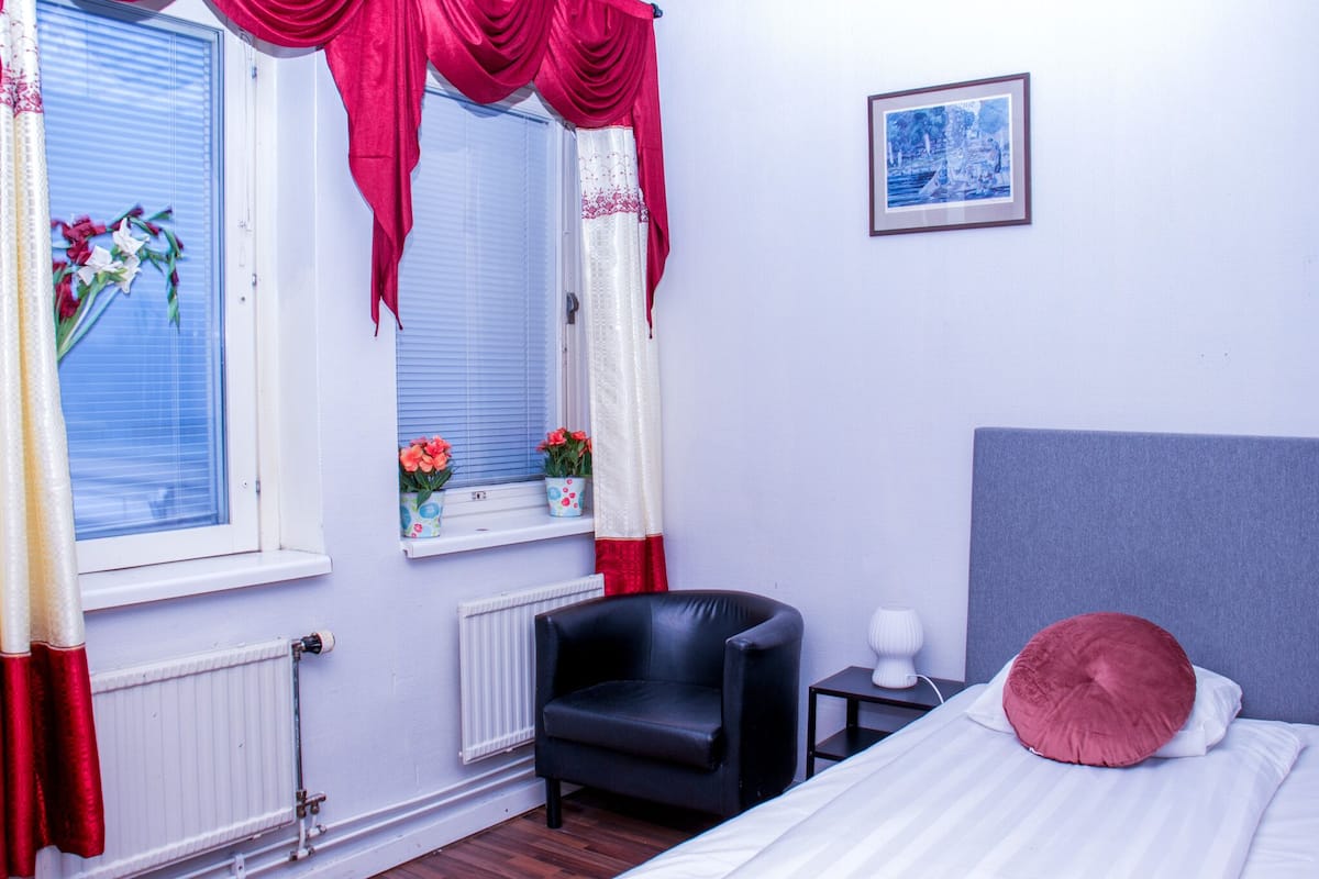 A bedroom with a bed, a black chair, a small table, a radiator, and a window with curtains.