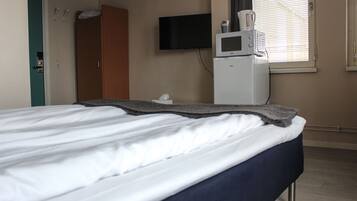 A hotel room with a bed, a television mounted on the wall, a microwave, and a window with blinds.