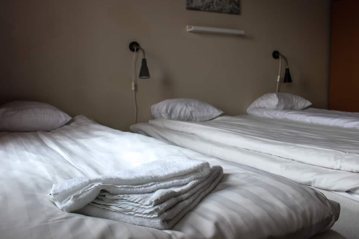 Two neatly made beds with white linens and pillows, a wall-mounted shelf above, and two wall-mounted lamps on either side of the beds.