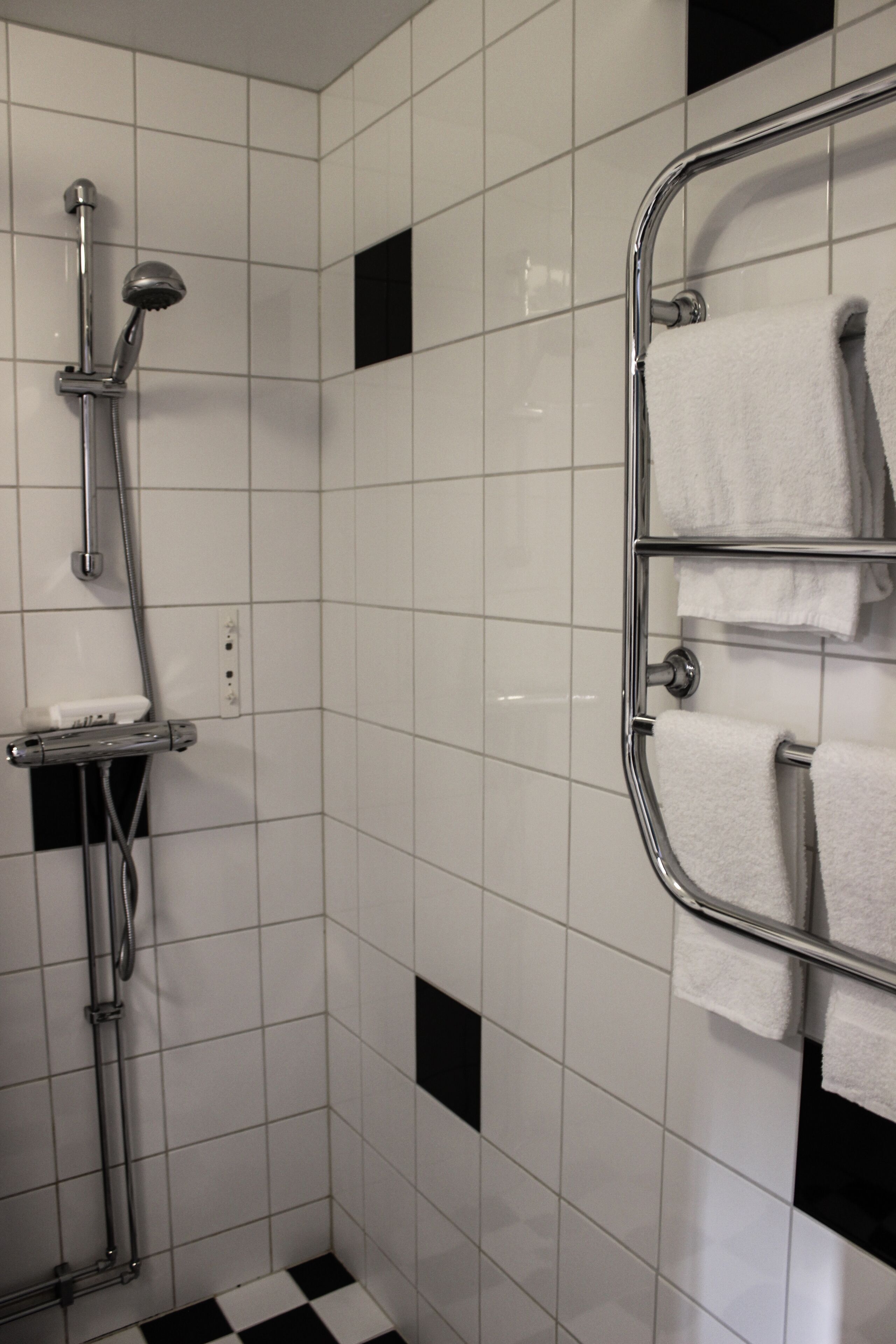 Un baño con paredes de azulejos blancos, suelo a cuadros blancos y negros, una ducha con manguera cromada y un perchero con varias toallas blancas.