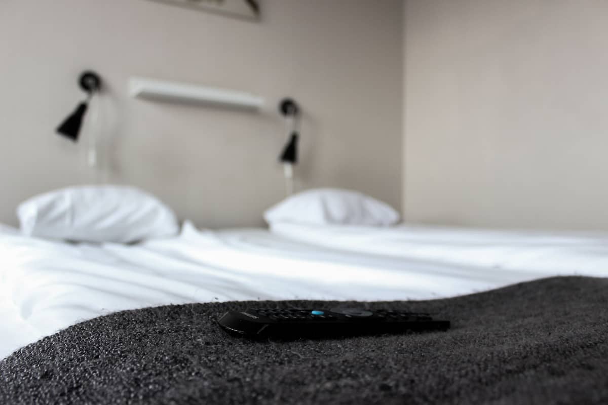 A bed with white sheets and pillows, a remote control on a dark surface, and a white bedside table with a lamp.