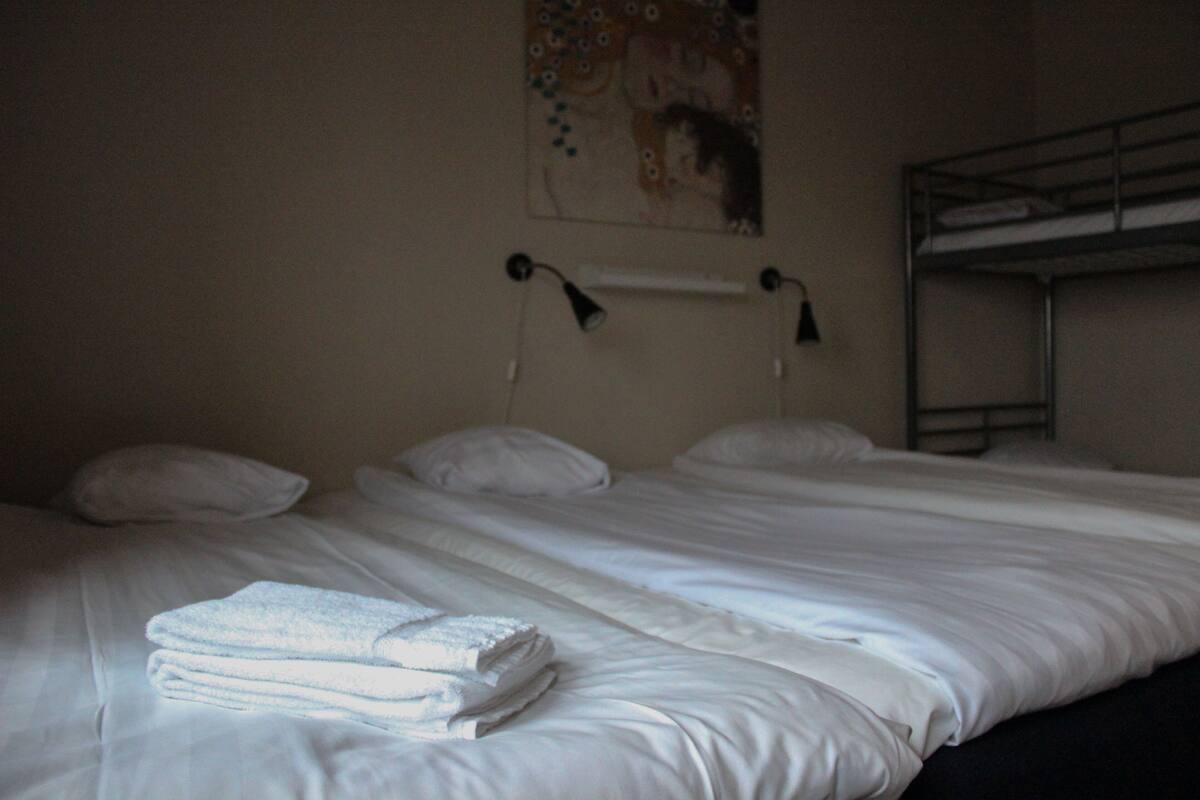 A single bed with white linens and a folded towel, a bedside lamp, and a bunk bed in the background.
