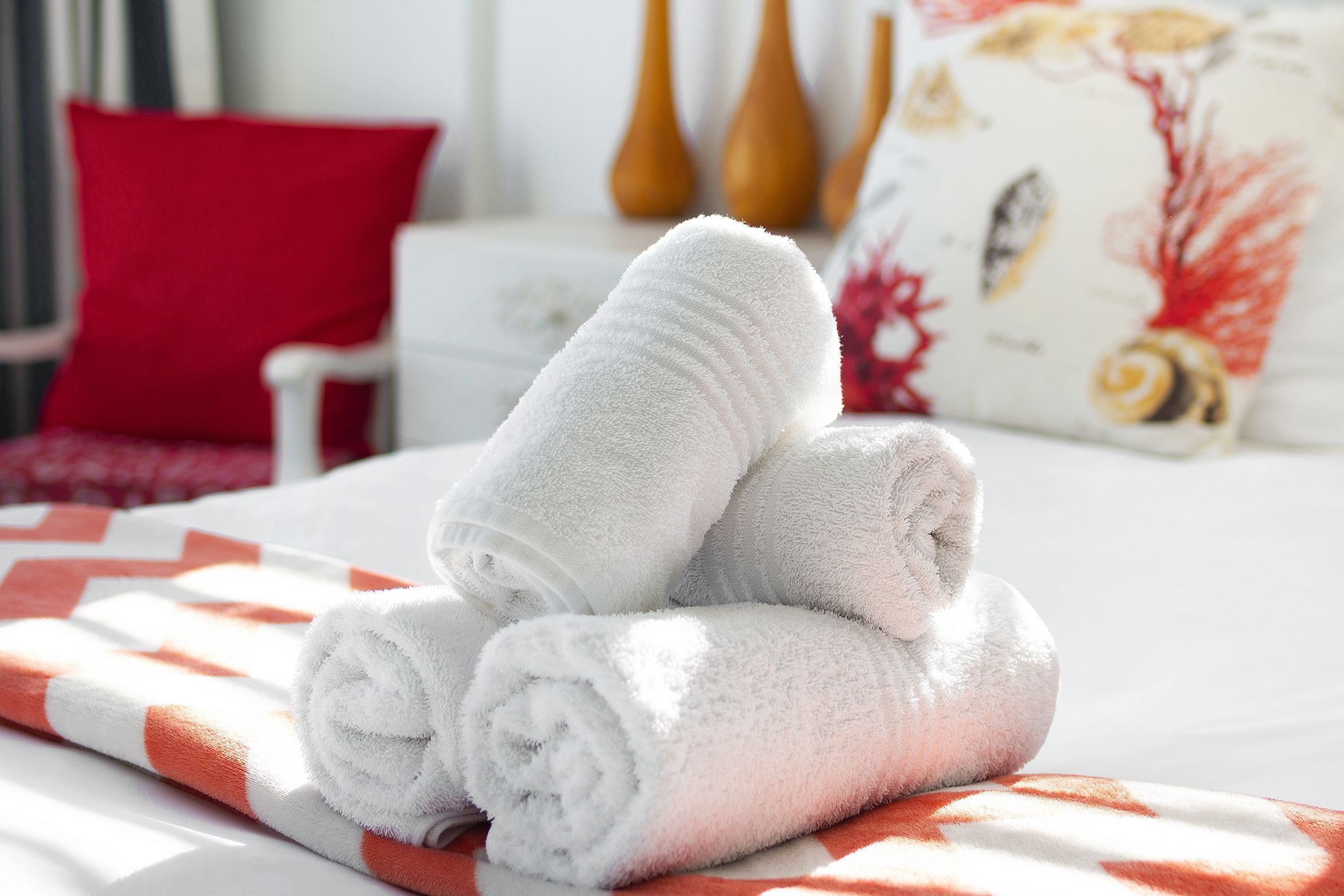 A neatly made bed with white towels folded on top, a red pillow, and a patterned pillow in the background.