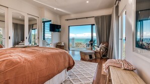 A spacious bedroom with a large bed, a wooden bench, and a view of the ocean through large windows. - Villa Sunshine - 200m to beach, ocean views, 5mins to Hastings St (Sunshine Beach)