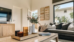 A modern living room with a wooden cabinet, a flat-screen TV, a coffee table with decorative items, a grey sofa with cushions, and a large window offering a view of the outdoors. - Villa Sunshine - 200m to beach, ocean views, 5mins to Hastings St (Sunshine Beach)