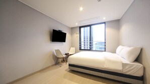 Una habitación de hotel moderna con una cama grande, un televisor de pantalla plana montado en la pared, un escritorio con una silla y una ventana con vistas a edificios.