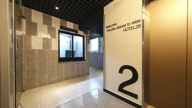 Un pasillo de hotel con una recepción, un letrero que dice 'Welcome. Ofrecemos espacios para relajarse HOTEL26', y el número '2' que indica la habitación.