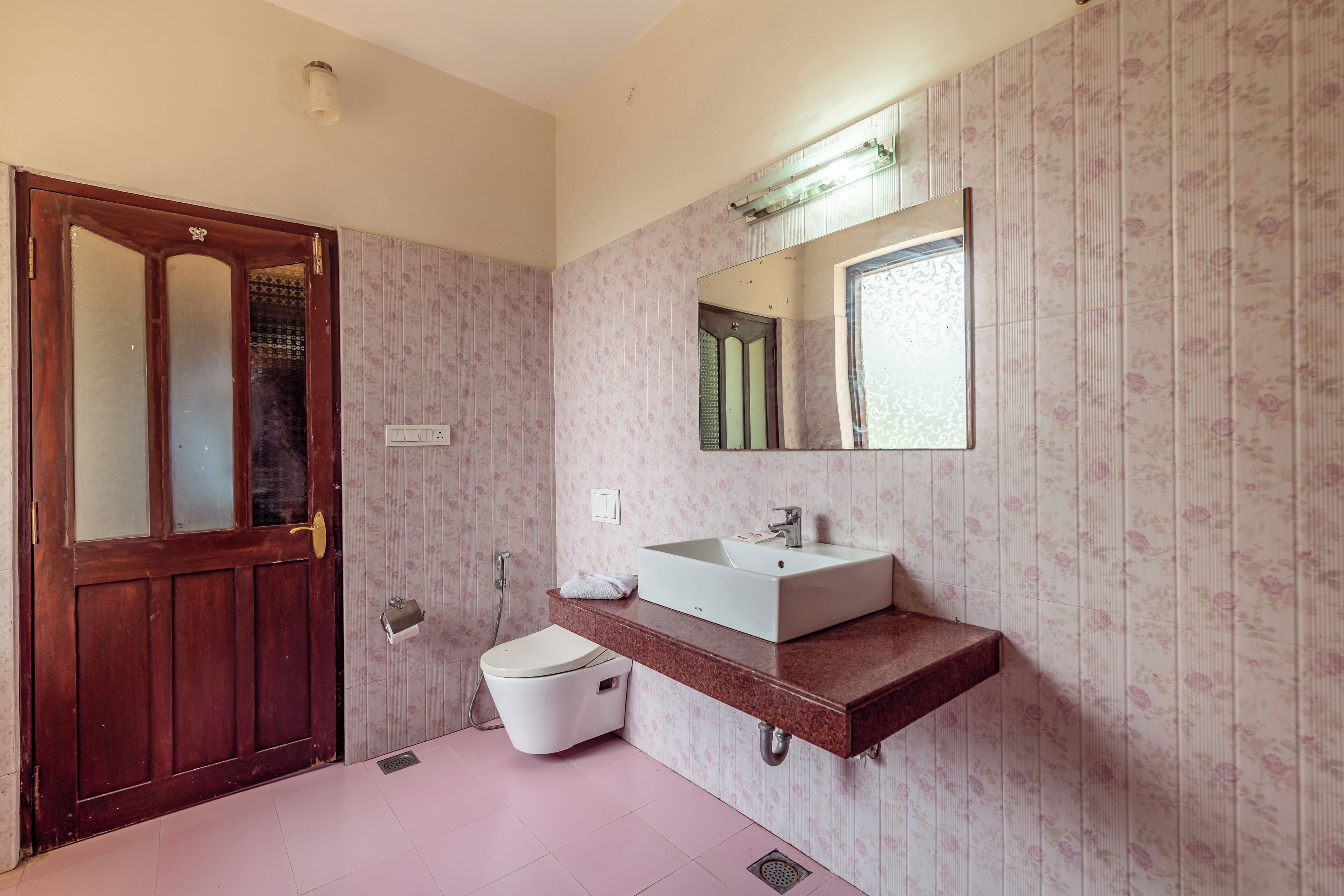 A bathroom with a wooden door, a white toilet, a sink with a mirror above it, and floral wallpaper.