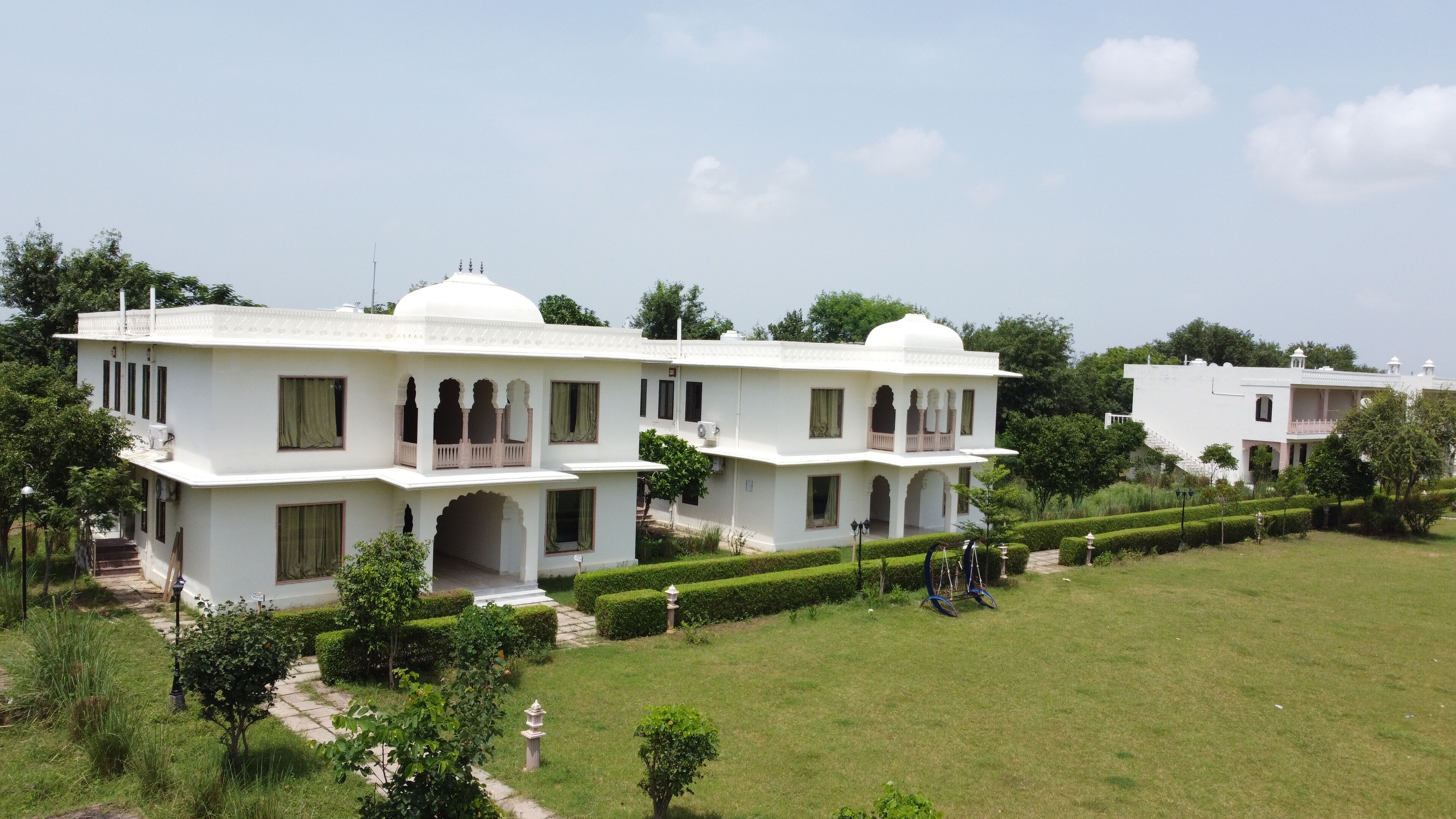 A white, two-story building with a flat roof, multiple balconies, and a garden with trimmed hedges.