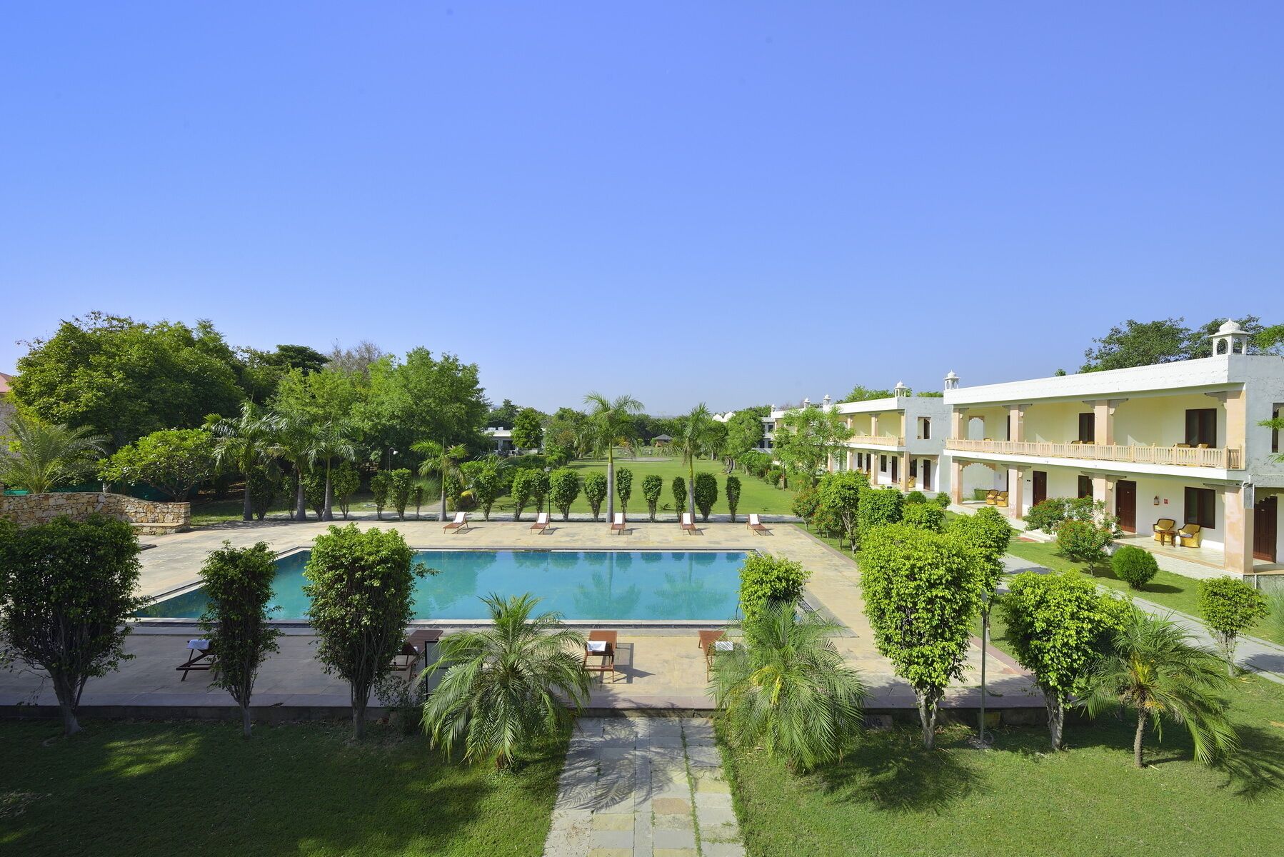 A pool surrounded by greenery, a building with multiple balconies, and a clear sky.