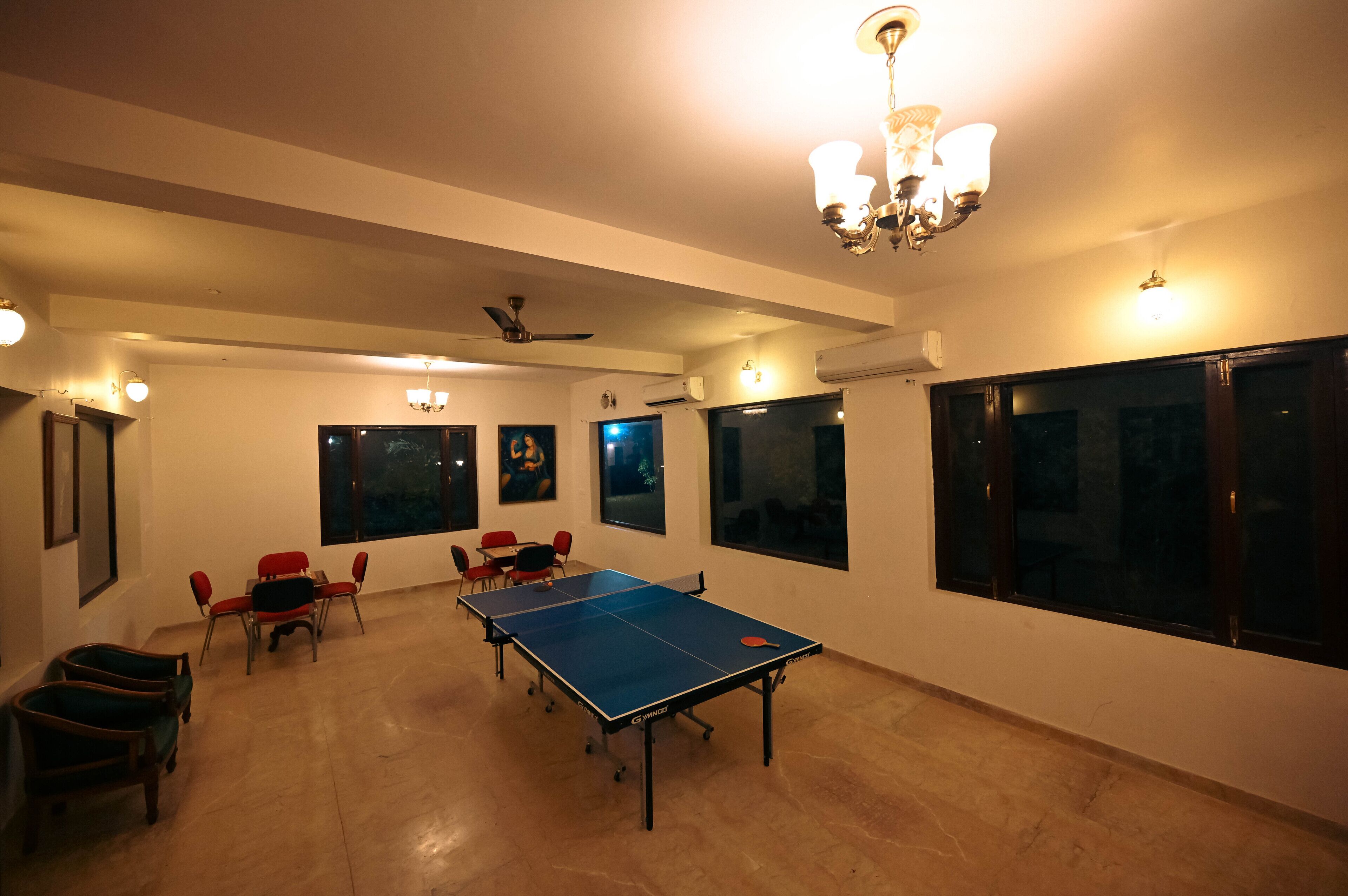 A room with a ping pong table, chairs, a ceiling fan, and large windows.