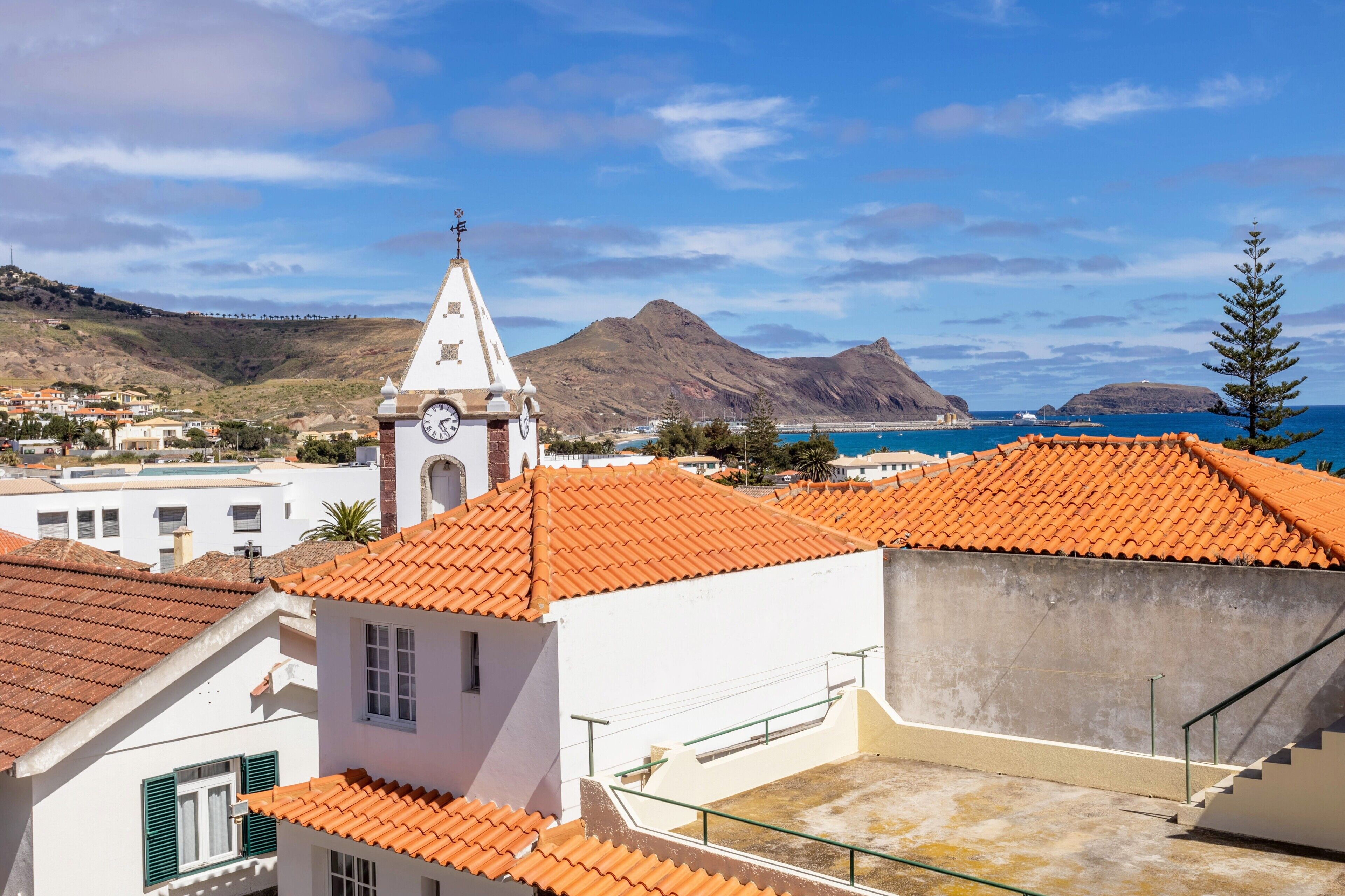 A coastal town with buildings featuring red-tiled roofs, a prominent clock tower, and a mountainous backdrop.