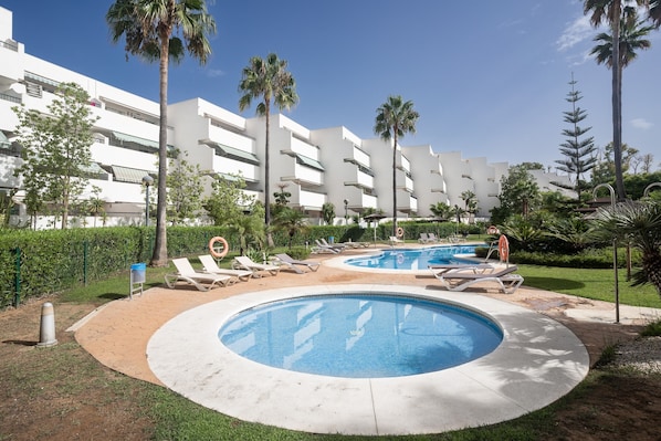 Zona de piscina con dos piscinas circulares, sillas de descanso, palmeras y un edificio moderno al fondo.