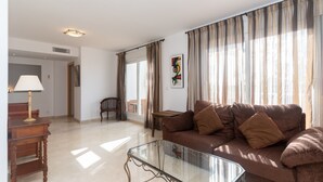 Ein Wohnzimmer mit Glastisch, braunem Sofa, Beistelltisch aus Holz und einer Lampe.