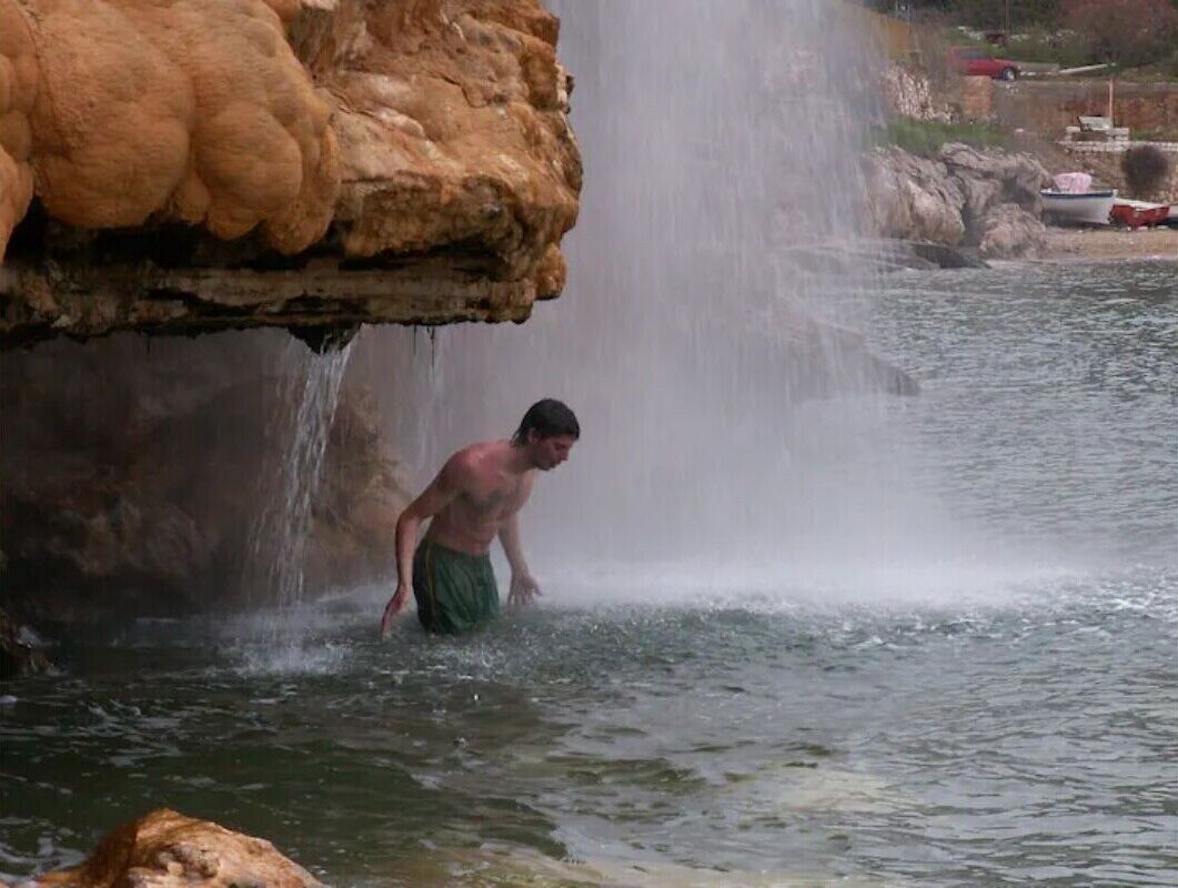A person in a natural setting with a waterfall and boats in the background.