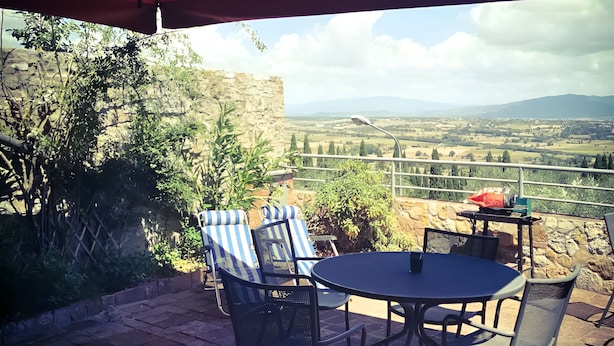 A patio with a table and chairs under a red umbrella, overlooking a scenic landscape with mountains and fields.